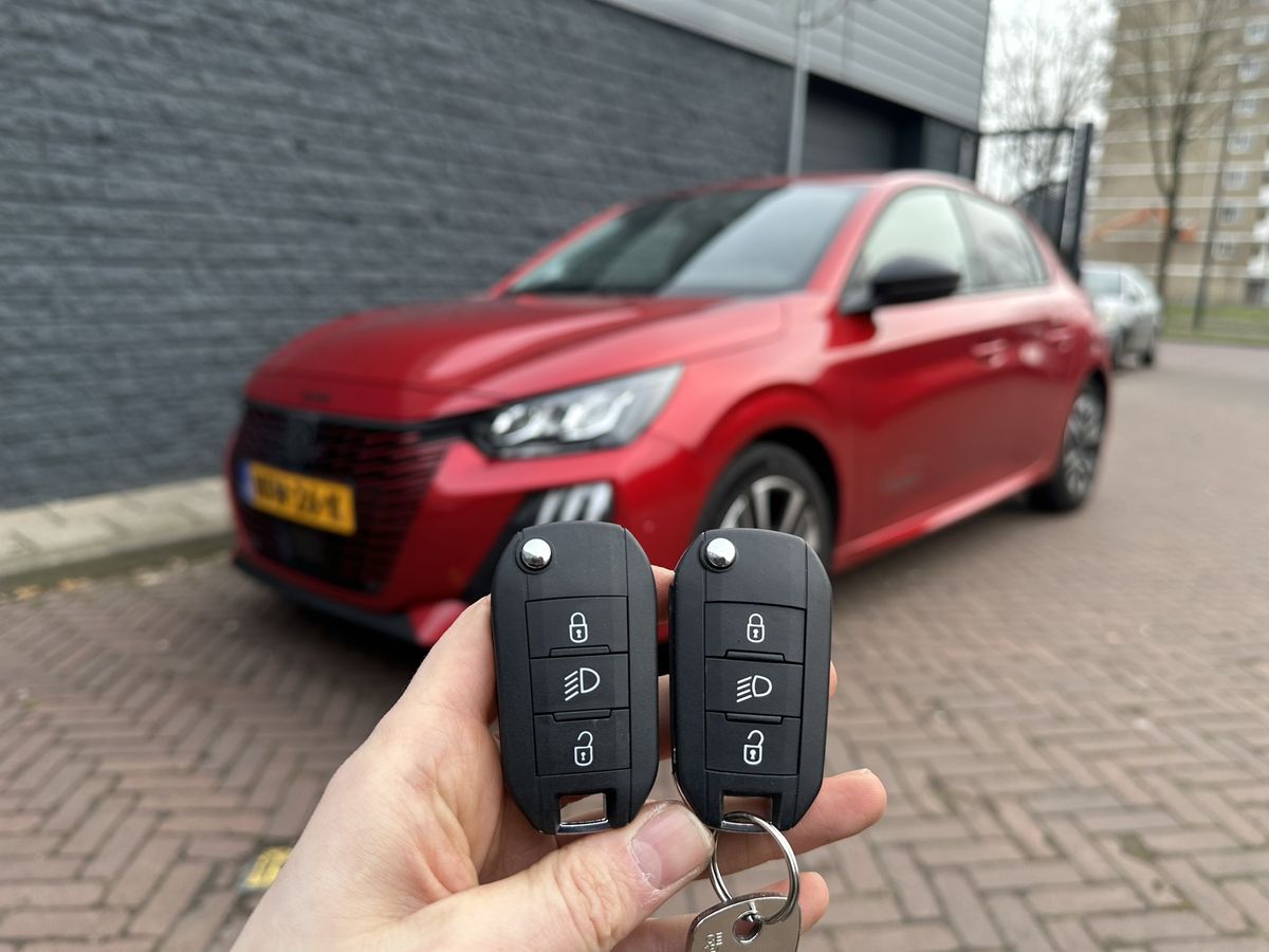 Twee Peugeot-klapsleutels in de hand, met een rode Peugeot 208 op de achtergrond